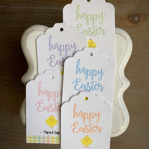 Puede incluir: Cuatro etiquetas de regalo blancas con bordes festoneados. Cada etiqueta presenta un pollito de color pastel diferente y el texto "happy Easter". Dos de las etiquetas tienen el texto "Papa & Gigi" en la parte inferior.
