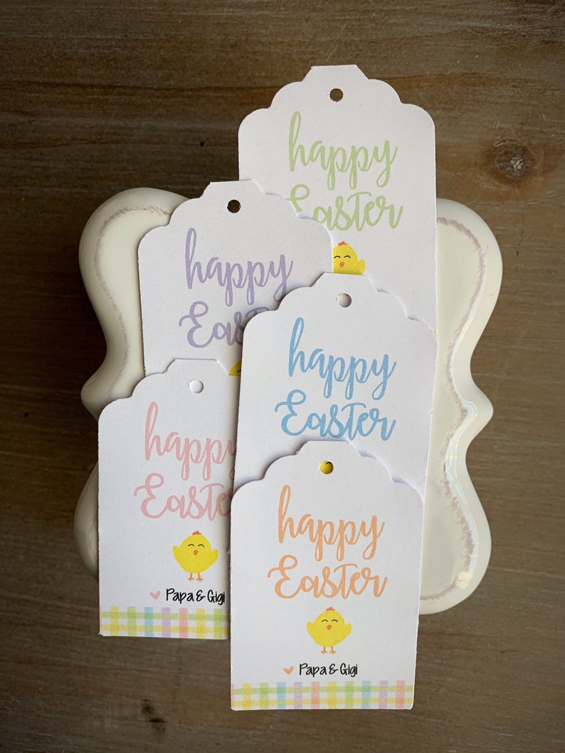 Puede incluir: Cuatro etiquetas de regalo blancas con bordes festoneados. Cada etiqueta presenta un pollito de color pastel diferente y el texto "happy Easter". Dos de las etiquetas tienen el texto "Papa & Gigi" en la parte inferior.