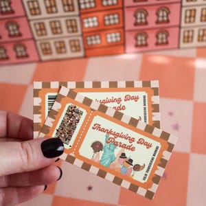 May include: Two rectangular paper tickets with a brown and white chequered border. The tickets feature the text Thanksgiving Day Parade, an illustration of the Statue of Liberty, and various balloons. One ticket includes a gold glitter accent.