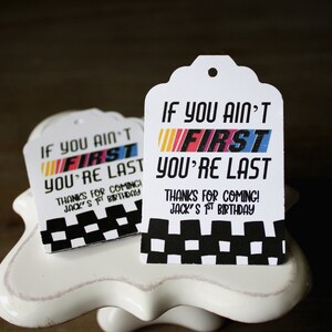 Puede incluir: Dos etiquetas de regalo blancas con bordes a cuadros en blanco y negro. Las etiquetas tienen el texto "If you ain't first you're last" con la palabra "first" en colores arco&iacute;ris. El texto "Thanks for coming! Jack's 1st birthday" est&aacute; impreso debajo del texto principal.