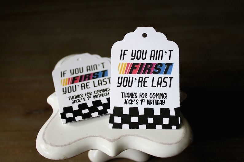 Puede incluir: Dos etiquetas de regalo blancas con bordes a cuadros en blanco y negro. Las etiquetas tienen el texto "If you ain't first you're last" con la palabra "first" en colores arco&iacute;ris. El texto "Thanks for coming! Jack's 1st birthday" est&aacute; impreso debajo del texto principal.