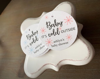 May include: Two round white gift tags with a snowflake design and the text "Baby it's cold outside" and "Ashlyn's baby shower".
