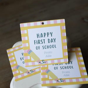 May include: Square paper gift tags featuring a yellow and pink checkered border. The centre text reads Happy First Day Of School with a yellow pencil illustration and the name Allie printed below. Each tag has a small hole at the top for hanging.