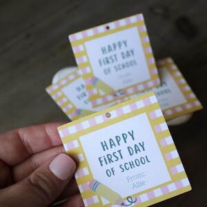 May include: A square paper gift tag with a yellow and white chequered border. The centre features the text "HAPPY FIRST DAY OF SCHOOL" in dark blue, with "From: Allie" below it and a small illustration of a yellow pencil with a pink eraser.