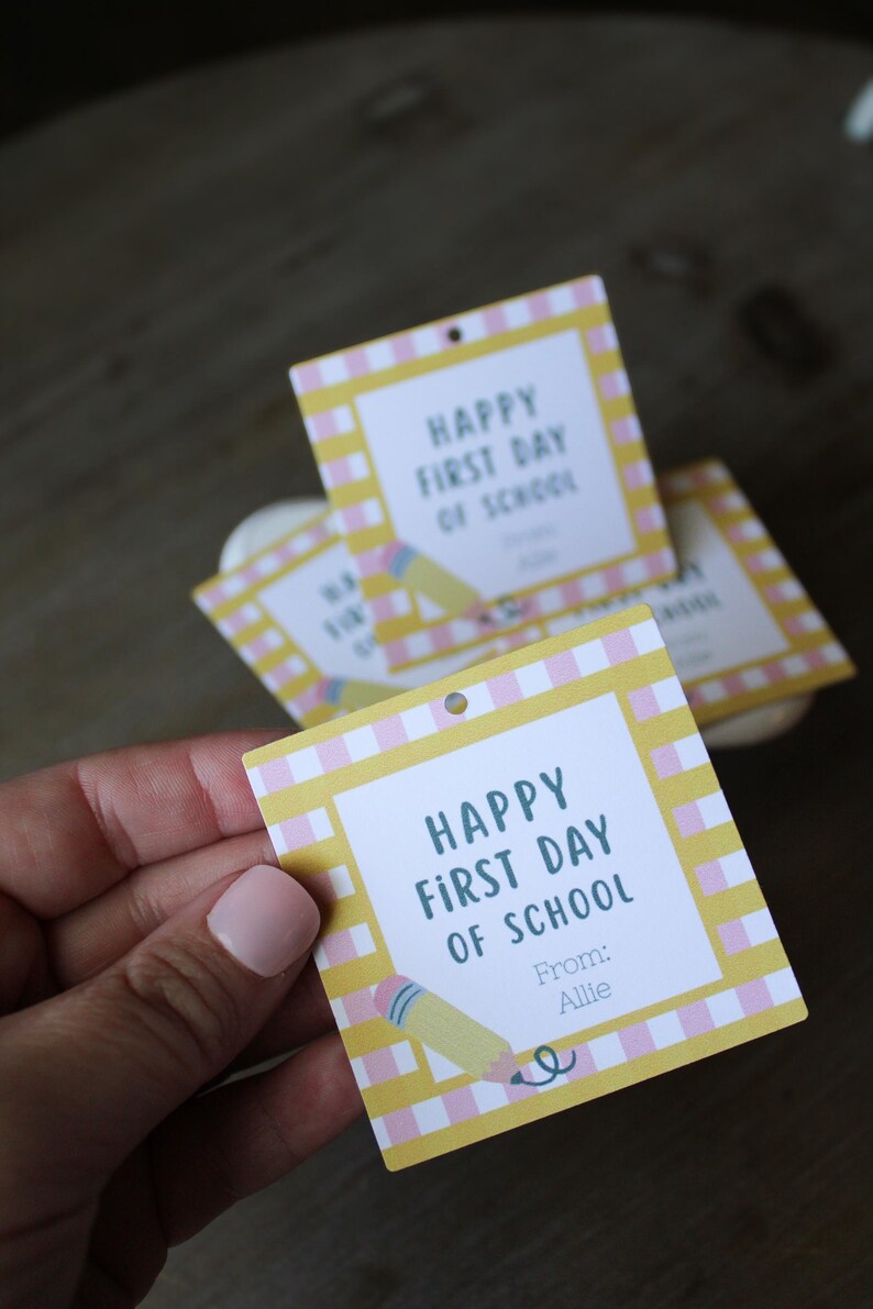 May include: A square paper gift tag with a yellow and white chequered border. The centre features the text "HAPPY FIRST DAY OF SCHOOL" in dark blue, with "From: Allie" below it and a small illustration of a yellow pencil with a pink eraser.