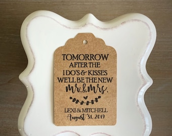 May include: A white, ornate, ceramic frame with a brown tag in the center. The tag reads "TOMORROW AFTER THE I DO'S & KISSES WE'LL BE THE NEW Mr.&Mrs. LEXI & MITCHELL August 31, 2019" in black text. The frame has a distressed, pinkish-brown trim.