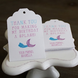 Puede incluir: Dos etiquetas blancas de agradecimiento con un diseño de cola de sirena. El texto en las etiquetas dice "¡Gracias por hacer de mi cumpleaños un splashi!" y "Tu amiga, Isabella".