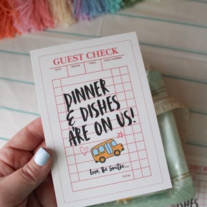 May include: A white rectangular guest check card featuring a red grid pattern and the text "GUEST CHECK" at the top. The card displays the black handwritten text "DINNER & DISHES ARE ON US!" above a small orange school bus illustration and "Love, The Smiths".