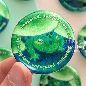 Peut inclure: Plusieurs boutons ronds verts avec un motif de grenouille. Les boutons présentent une illustration de grenouille de dessin animé avec le texte « bothered, dehydrated, sad, not in my lane, unfocused, withering ». Le logo Citrus Sailing est également visible.