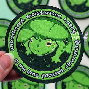 May include: A green embroidered patch with a cartoon frog design. The patch includes the words "unbothered, moisturised, happy, in my lane, focused, flourishing" in a circular layout. Other similar patches are visible in the background.