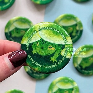 Könnte beinhalten: Grüner Frosch-Button mit glitzernder Oberfläche. Der Frosch sitzt unter einem Blatt und der Text um ihn herum lautet "unbothered, moisturised, happy. In my lane. Focused, flourishing."