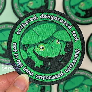 May include: A circular green patch with a cartoon frog design and the words "bothered, dehydrated, sad, not in my lane, unfocused, withering." The frog is green with large eyes and is inside a green circle.
