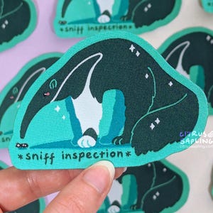 May include: A teal sticker with a cartoon anteater in dark green, white, and teal. The anteater is sniffing the ground, with the words "*sniff inspection*" below. The sticker has a textured appearance and a small ant is visible.