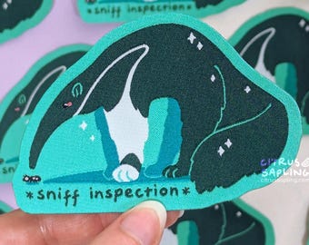 May include: A teal sticker with a cartoon anteater in dark green, white, and teal. The anteater is sniffing the ground, with the words "*sniff inspection*" below. The sticker has a textured appearance and a small ant is visible.