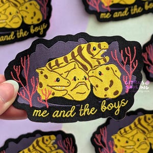 Könnte beinhalten: Gestickter Aufnäher mit einer Gruppe gelber, aalartiger Kreaturen mit braunen Streifen und Flecken, vor einem lila Hintergrund mit Korallenakzenten. Der Text "me and the boys" ist in Gold gestickt.