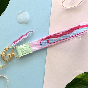 Op de afbeelding: Een stoffen polsband-sleutelhanger met een roze en lichtblauw patroon met een stripfiguur, bevestigd aan een goudkleurige karabijnhaak en sleutelring. Een kleine, doorzichtige acryl bedel met een bijpassend ontwerp is ook bevestigd.