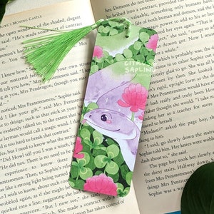 May include: A bookmark featuring a watercolour illustration of a purple snake with pink eyes, curled around green clover leaves and pink flowers. The bookmark has a green tassel.