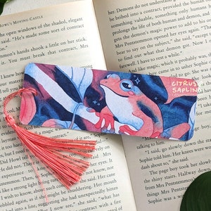 Könnte beinhalten: Ein pinkfarbener Frosch-Lesezeichen mit blauem und weißem Hintergrund. Das Lesezeichen hat einen Quasten und den Text "CITRUS SAPLING" unten.