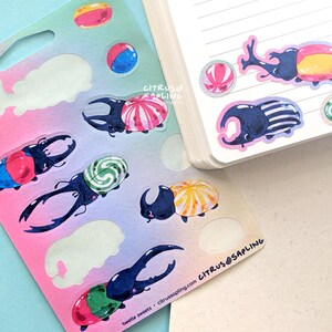 May include: A sheet of colorful stickers featuring illustrated beetles with candy-like shells and various candies. The stickers are on a pink and blue gradient background. The text "beetle sweets" and "citrus@sapling.com" is visible.