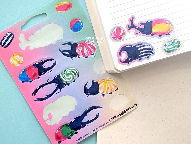 May include: A sheet of colorful stickers featuring illustrated beetles with candy-like shells and various candies. The stickers are on a pink and blue gradient background. The text "beetle sweets" and "citrus@sapling.com" is visible.