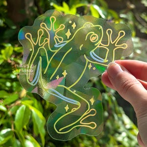 Puede incluir: Una pegatina holográfica con un diseño de rana estilizada con contornos y acentos amarillos. La pegatina tiene un fondo transparente y se sostiene contra un fondo de follaje verde. La pegatina tiene el texto "Citrus Sapling".