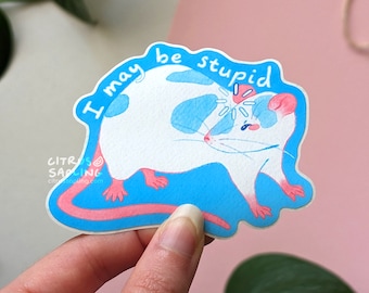 Op de afbeelding: Een gestanste sticker met een witte rat met blauwe vlekken en roze accenten. De rat heeft een bedroefde uitdrukking, met de tekst "I may be stupid" in een blauwe rand. De sticker is ontworpen in aquarel stijl.