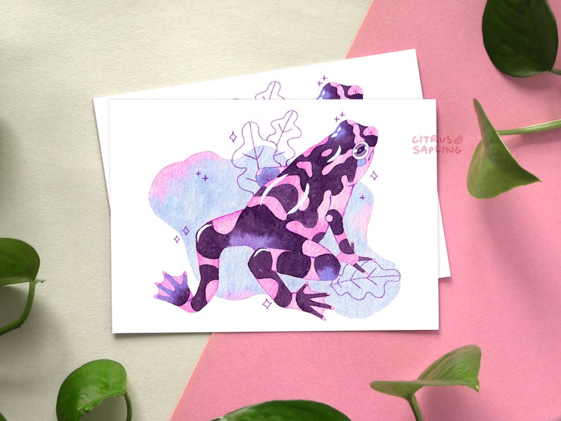 May include: A watercolor illustration of a pink and purple frog with black spots sitting on a leaf. The frog is surrounded by a blue and pink watercolor background with white stars.