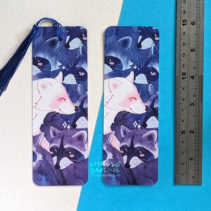 May include: Two watercolor bookmarks featuring a white fox and raccoons in a purple and blue color scheme. The bookmarks are printed with the text "CITRUS SAPLING" and "citrussapling.com".