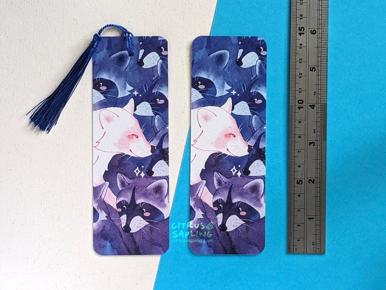 May include: Two watercolor bookmarks featuring a white fox and raccoons in a purple and blue color scheme. The bookmarks are printed with the text "CITRUS SAPLING" and "citrussapling.com".