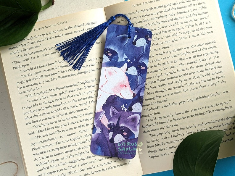 May include: A watercolor bookmark featuring a white fox and three raccoons with purple and blue accents. The bookmark has a blue tassel and the text "Citrus Sapling" in white.