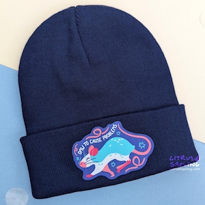 Puede incluir: Gorro de punto azul marino con un parche que presenta una rata de dibujos animados y el texto "Only to Cause Problems". La rata es de color azul claro y blanco con detalles en rosa. El gorro es de punto suave.
