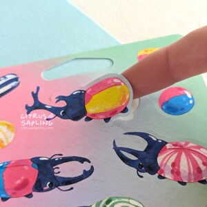 May include: A sheet of colorful stickers featuring cartoon beetles with various designs, including pink, blue, and yellow accents. The stickers are being held by a finger. The Citrus Sapling logo is visible.