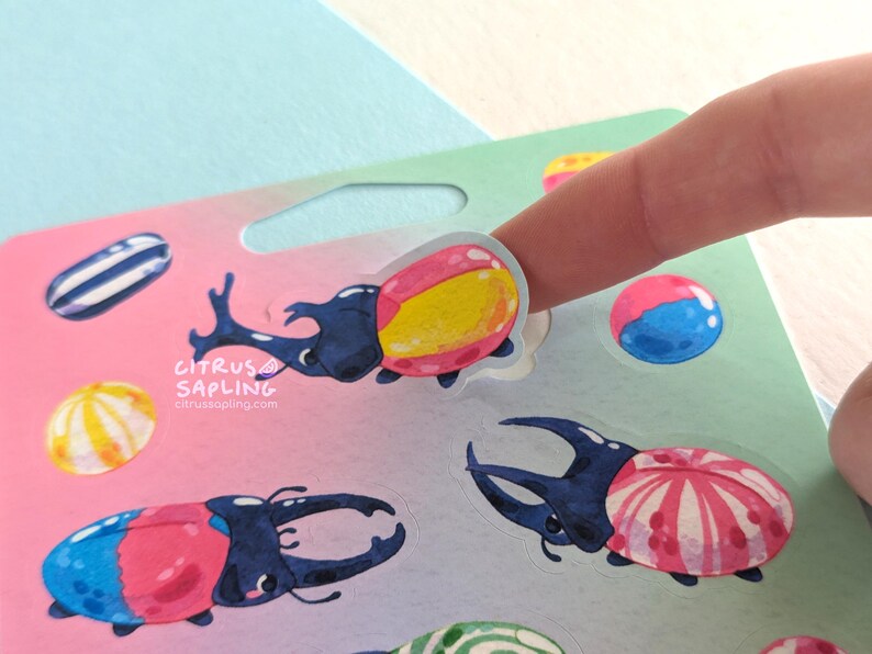 May include: A sheet of colorful stickers featuring cartoon beetles with various designs, including pink, blue, and yellow accents. The stickers are being held by a finger. The Citrus Sapling logo is visible.