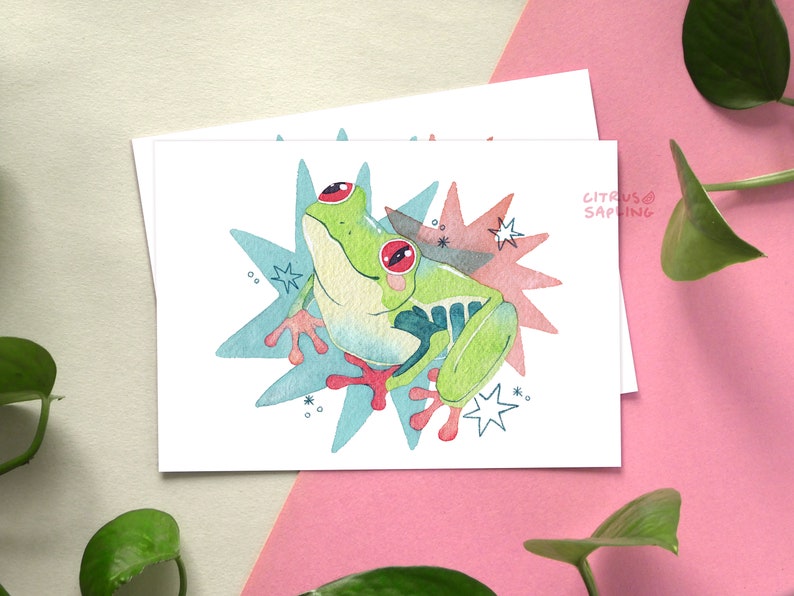 May include: A watercolor illustration of a green tree frog with red eyes, sitting on a blue and pink starburst background. The text "CITRUS SAPLING" is written in the top right corner.