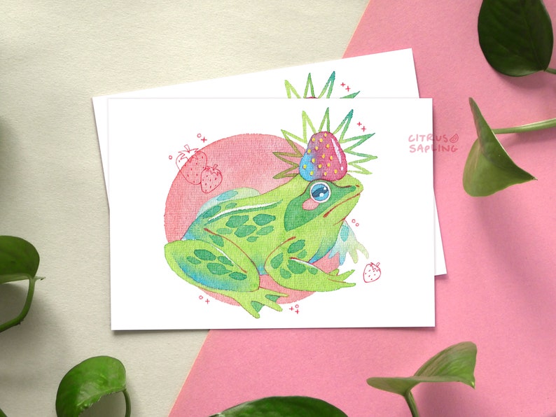 May include: A green frog with a pink strawberry crown sits on a pink and white background. The frog is surrounded by small pink strawberries.