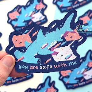 May include: A dark blue sticker featuring a light blue hammerhead shark surrounded by pink stingrays and small fish. The text "you are safe with me" is printed in white and pink letters at the bottom. The design includes small bubbles.