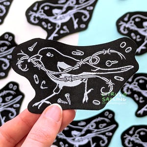 May include: A black embroidered patch featuring a white bird silhouette holding a piece of barbed wire in its beak. The design includes scattered small metal objects like screws, paperclips, and washers. The text Citrus Sapling is visible.