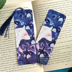 May include: Two blue and purple bookmarks with a watercolor illustration of raccoons and foxes. The bookmarks feature a white fox with pink accents and a purple raccoon with black accents. The bookmarks are set against a dark blue background with white stars.