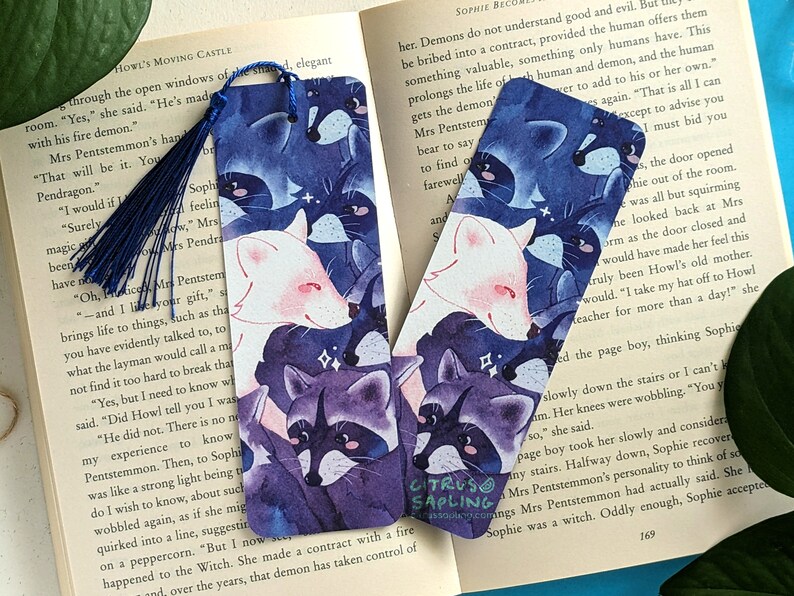 May include: Two blue and purple bookmarks with a watercolor illustration of raccoons and foxes. The bookmarks feature a white fox with pink accents and a purple raccoon with black accents. The bookmarks are set against a dark blue background with white stars.