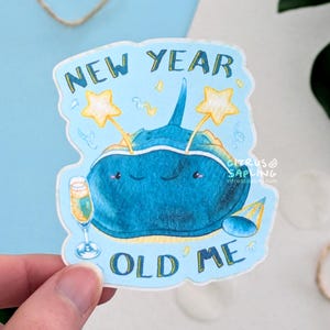 May include: A sticker with a blue stingray illustration, featuring the text "NEW YEAR OLD ME." The stingray has a smiling face and is decorated with star-shaped wands, a champagne glass, and a party hat. The sticker is held by a hand.