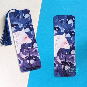 May include: A blue and purple watercolor bookmark featuring a white fox and raccoons with sparkling details. The bookmark has a blue tassel and the text "CITRUS SAPLING" at the bottom.