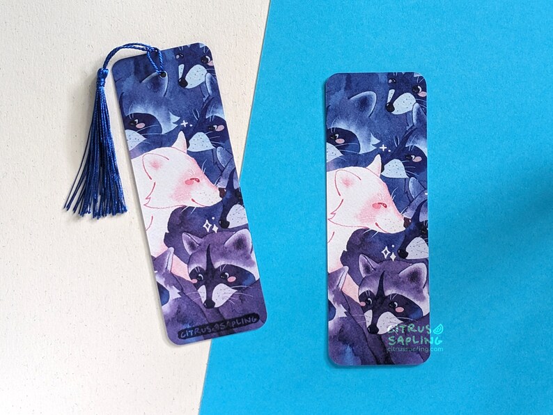May include: A blue and purple watercolor bookmark featuring a white fox and raccoons with sparkling details. The bookmark has a blue tassel and the text "CITRUS SAPLING" at the bottom.