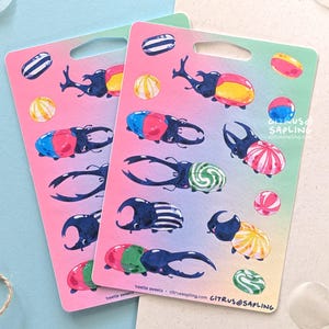 May include: Two sticker sheets with colorful illustrations of beetles and candies. The beetles are depicted with various patterns and colors, and the candies are round and striped. The background is a gradient of pink and green.
