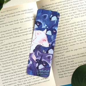 May include: A watercolor bookmark featuring a white fox, a raccoon, and a black cat. The animals are drawn in a whimsical style with a blue and purple background. The bookmark has the text "Citrus Sapling" in the bottom right corner.