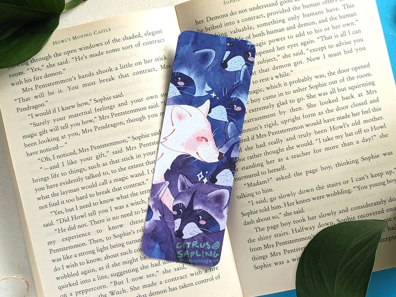 May include: A watercolor bookmark featuring a white fox, a raccoon, and a black cat. The animals are drawn in a whimsical style with a blue and purple background. The bookmark has the text "Citrus Sapling" in the bottom right corner.