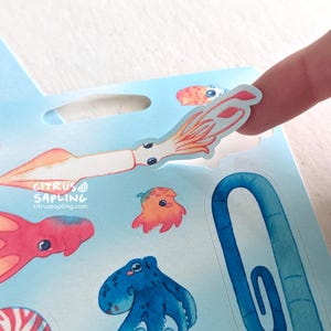 May include: A sheet of colorful ocean-themed stickers featuring various sea creatures, including a squid, octopus, and jellyfish. A finger is peeling off a squid sticker. The stickers have a watercolor style.