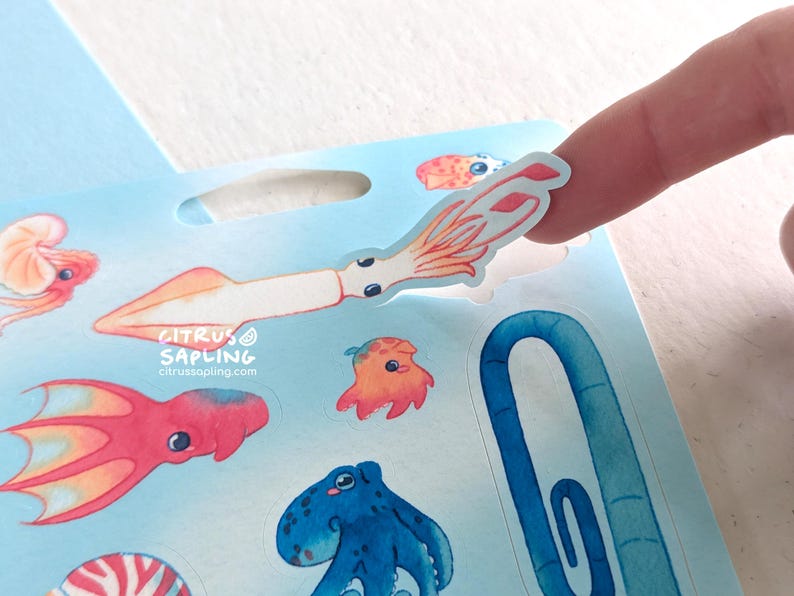 May include: A sheet of colorful ocean-themed stickers featuring various sea creatures, including a squid, octopus, and jellyfish. A finger is peeling off a squid sticker. The stickers have a watercolor style.