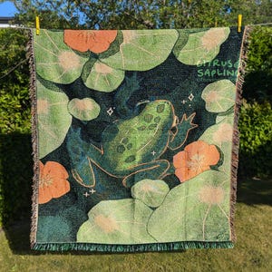 Puede incluir: Una manta tejida con un diseño de rana. La manta muestra una rana verde entre nenúfares y flores naranjas, con el texto "Citrus Sapling". La paleta de colores incluye tonos de verde, naranja y azul oscuro.