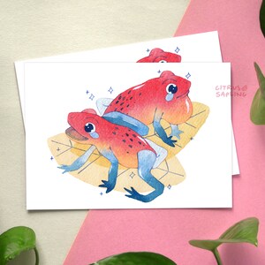 May include: Two red and orange poison dart frogs with blue accents are sitting on yellow leaves with white sparkles. The frogs are facing each other and have a sad expression. The text "CITRUS SAPLING" is in the top right corner of the image.