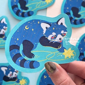 Puede incluir: Un parche bordado azul y negro de un panda rojo sosteniendo una estrella amarilla. El panda está rodeado de un cielo estrellado azul y blanco. El parche está sobre un fondo azul claro.